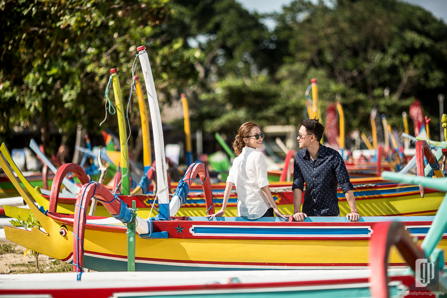 casual prewedding in sanur bali daylight beach with blue sky and traditional boats sunglasses smile happy couple